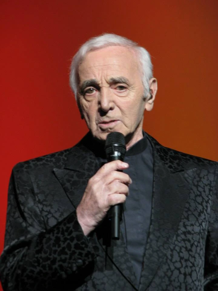Charles Aznavour holding a microphone on stage in front of a red background
