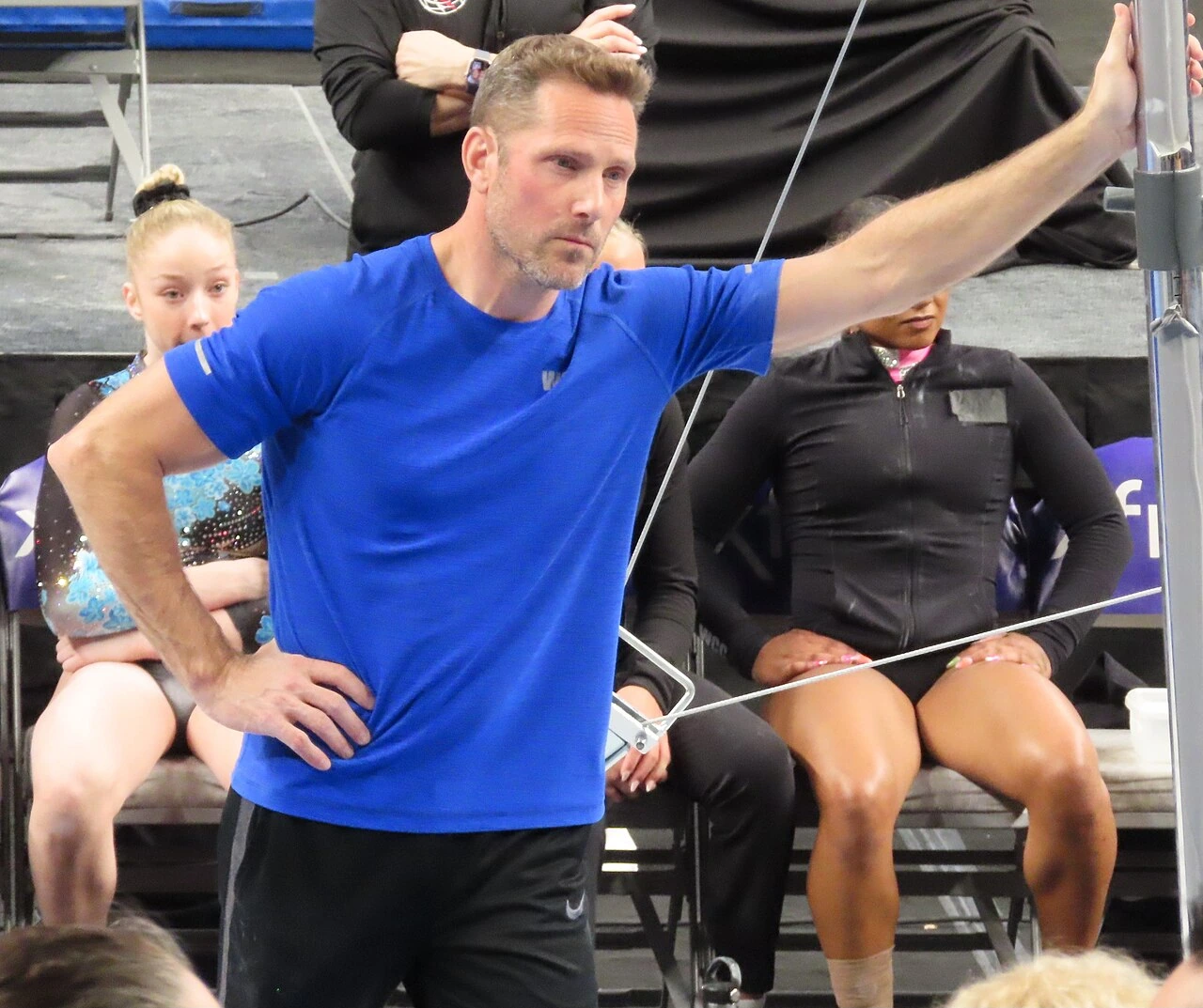 Laurent Landi at a gymnastics venue, wearing a blue shirt and gesturing while observing practice.