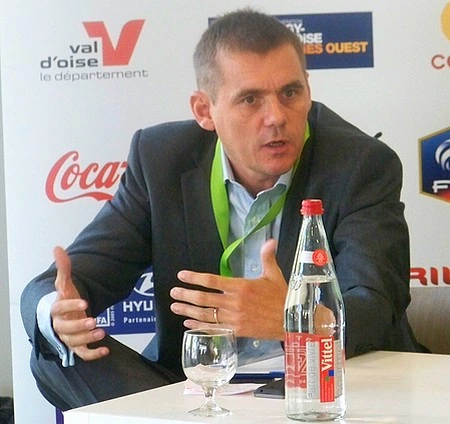 Olivier Leclercq speaking at an event, seated at a table with a microphone and sponsor backdrop behind him.